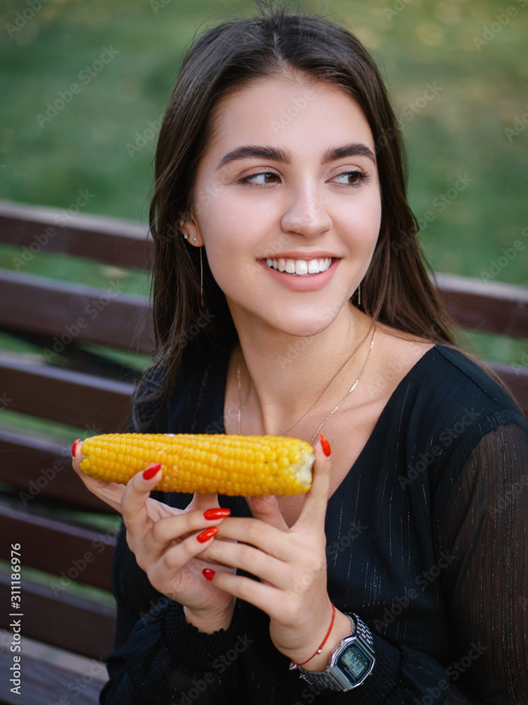 Outdoor summer lifestyle portrait of young beautiful brunette woman in black dress sitting on bench city park holding baked corn and adorable smiling Outdoor summer lifestyle portrait of young beautiful brunette woman in black dress sitting on bench city park holding baked corn and adorable smiling