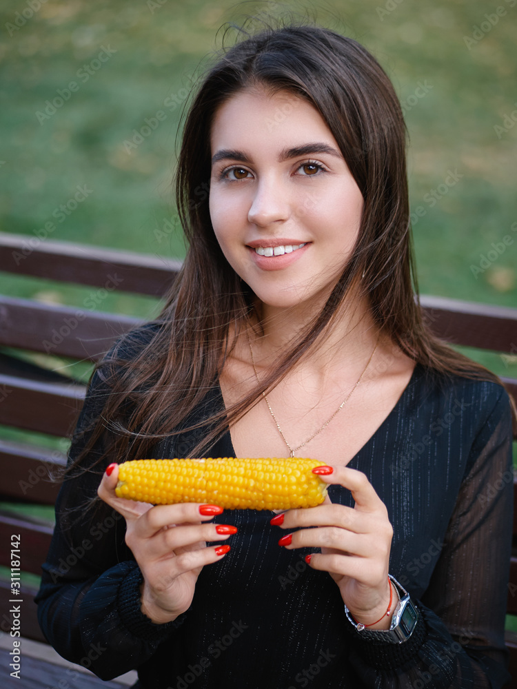 Outdoor summer lifestyle portrait of young beautiful brunette woman in black dress sitting on bench city park holding baked corn and adorable smiling Outdoor summer lifestyle portrait of young beautiful brunette woman in black dress sitting on bench city park holding baked corn and adorable smiling
