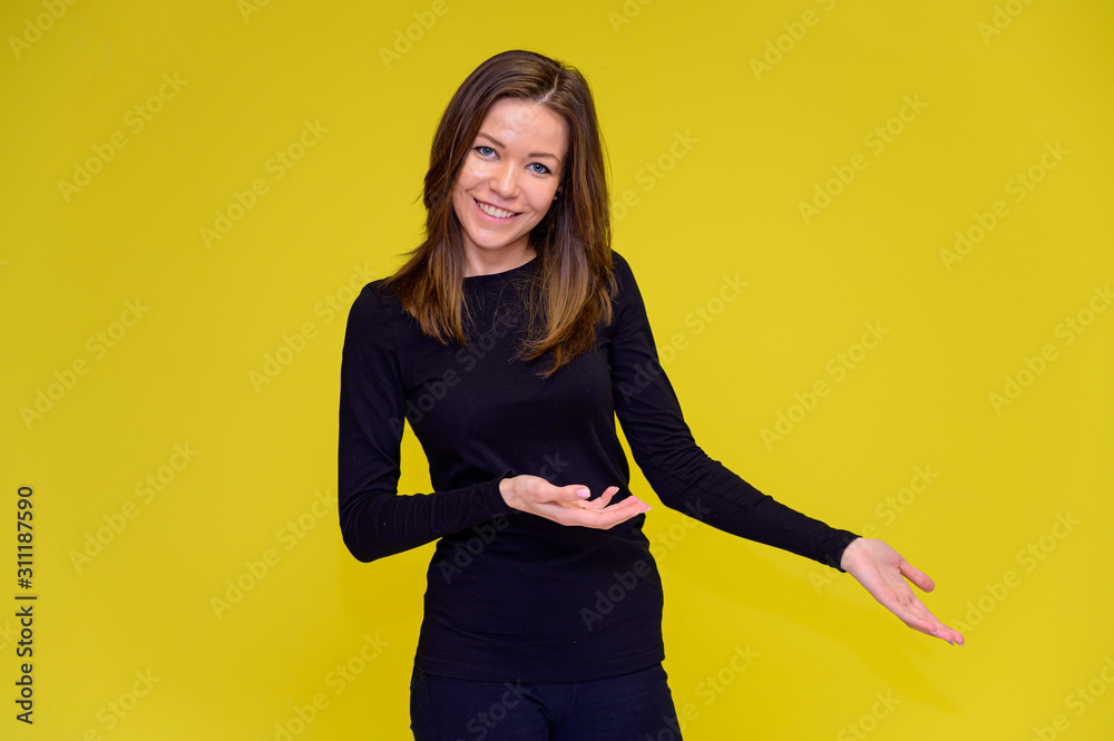 The concept of a smiling cute girl with positive emotions. Portrait of a young woman in a black T-shirt stands in different poses with long hair on a yellow background. Shows hands, talking. The concept of a smiling cute girl with positive emotions. Portrait of a young woman in a black T-shirt stands in different poses with long hair on a yellow background. Shows hands, talking.