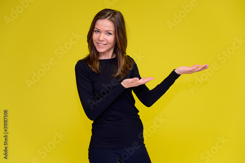The concept of a smiling cute girl with positive emotions. Portrait of a young woman in a black T-shirt stands in different poses with long hair on a yellow background. Shows hands, talking.