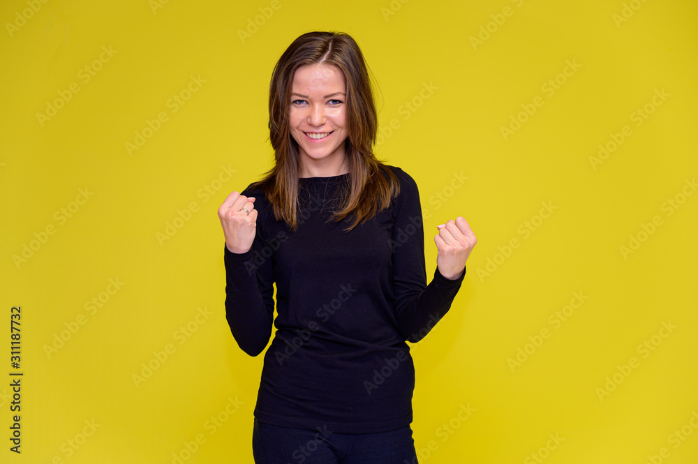 The concept of a smiling cute girl with positive emotions. Portrait of a young woman in a black T-shirt stands in different poses with long hair on a yellow background. Shows hands, talking. The concept of a smiling cute girl with positive emotions. Portrait of a young woman in a black T-shirt stands in different poses with long hair on a yellow background. Shows hands, talking.