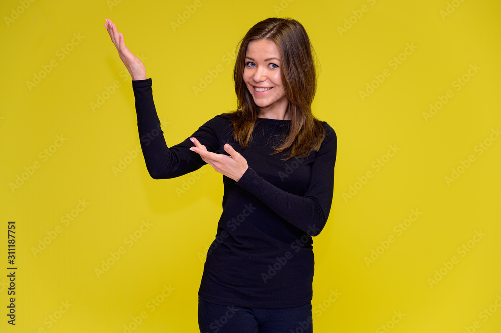 The concept of a smiling cute girl with positive emotions. Portrait of a young woman in a black T-shirt stands in different poses with long hair on a yellow background. Shows hands, talking. The concept of a smiling cute girl with positive emotions. Portrait of a young woman in a black T-shirt stands in different poses with long hair on a yellow background. Shows hands, talking.