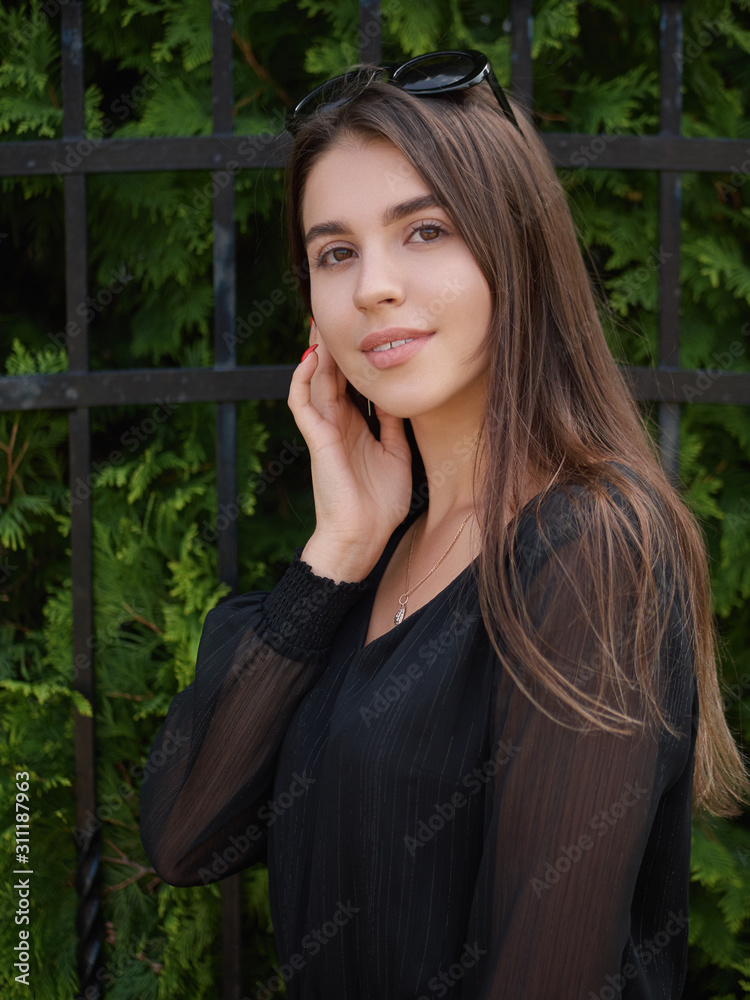 Outdoor summer lifestyle portrait of young beautiful brunette woman in black dress enjoying warm weather posing near fence city park with adorable smiling touching face Outdoor summer lifestyle portrait of young beautiful brunette woman in black dress enjoying warm weather posing near fence city park with adorable smiling touching face