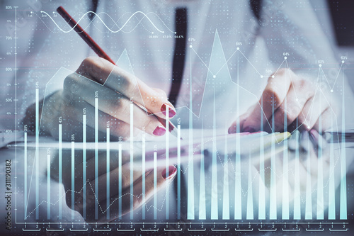 A woman hands writing information about stock market in notepad. Forex chart holograms in front. Concept of research.