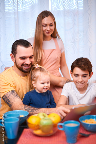 lovely adorable happy caucasian family consisted of young mother with long hair, bearded father and little kids son and daughter sit together on table, using laptop and eating, having meal, breakfast