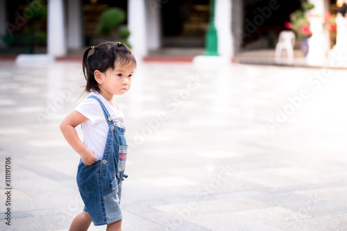 Side view of cute Asian little girl walking around the courtyard of Wat Sutad. A 2 years old in a blue denim overalls. Concept of taking children on a trip. © Kanthita