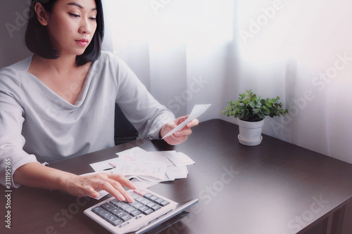 Asian woman hold receipt, slip, report or statement and press calculator with feeling stress, unhappy, worry about the calculate of prepare money for payment to burden of over shopping of credit card.