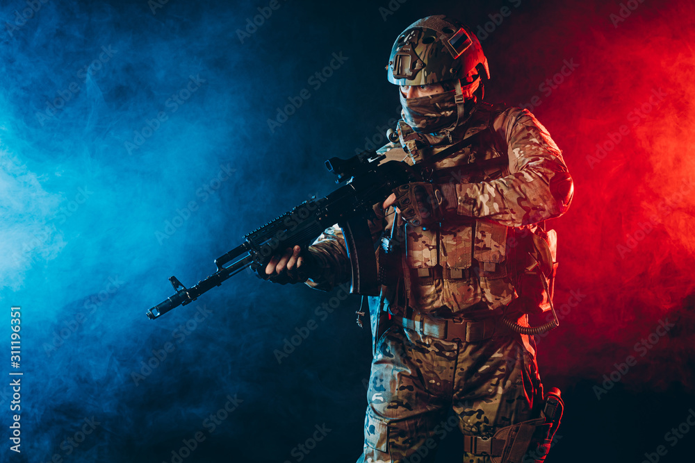 Naklejka premium safety and protection concept. young man work as military man protecting country, wearing military forces clothes and holding gun in smoky UV space