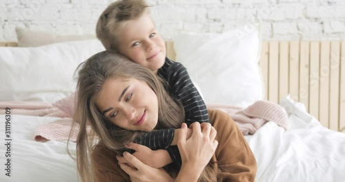Motherhood and childhood. Portrait of young woman mother and boy at home. Happy mom and her little son cuddling in bedroom looking at camera. Family lovely moments. Love and care of each other.