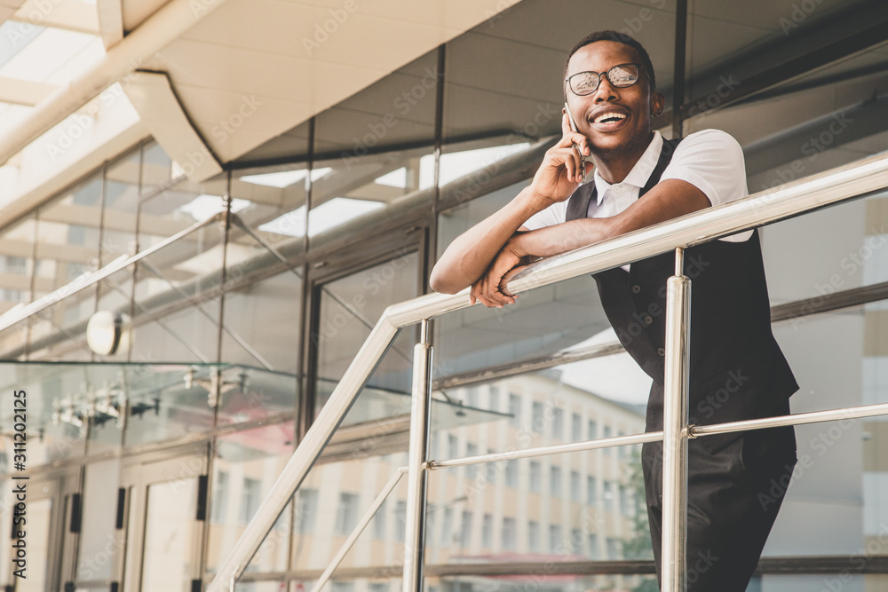 Young african american business man in suit and eyeglasses talking on the phone on the background of the business center Young african american business man in suit and eyeglasses talking on the phone on the background of the business center