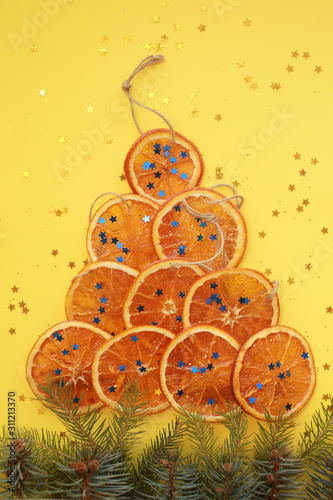 Ten dried orange slices with cords, imitating christmas tree with pine tree branches and blue glitter stars on yellow background