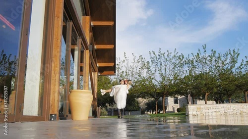 Handsome hispanic groom lifting and carrying blonde caucasian bride along grand wooden terrace.