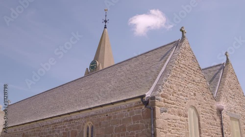 A daylight low angle shot of the pitched rood and spire of a church?s clock tower with exterior walls made of stone while the clear blue sky can be seen in the background...