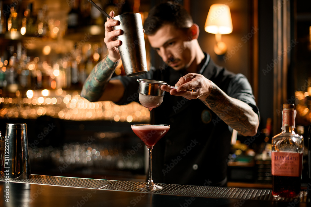 Tattooed bartender pouring a alcoholic drink from the steel shaker to the glass through the sieve