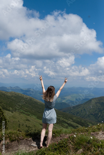A young woman in a blue dress looks up at the mountains from above, her hands raised