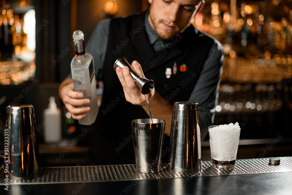 Male bartender pouring a vodka from the jigger to a steel shaker holding a bottle