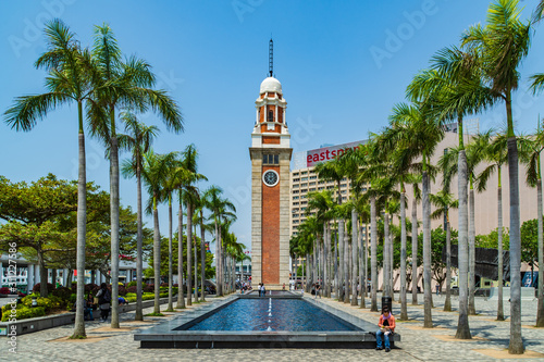 A man sitting on the fountain surrounded by trees in front of the Former Kowloon-Canton Railway Clock Tower in Tsim Sha Tsui, Hong Kong.