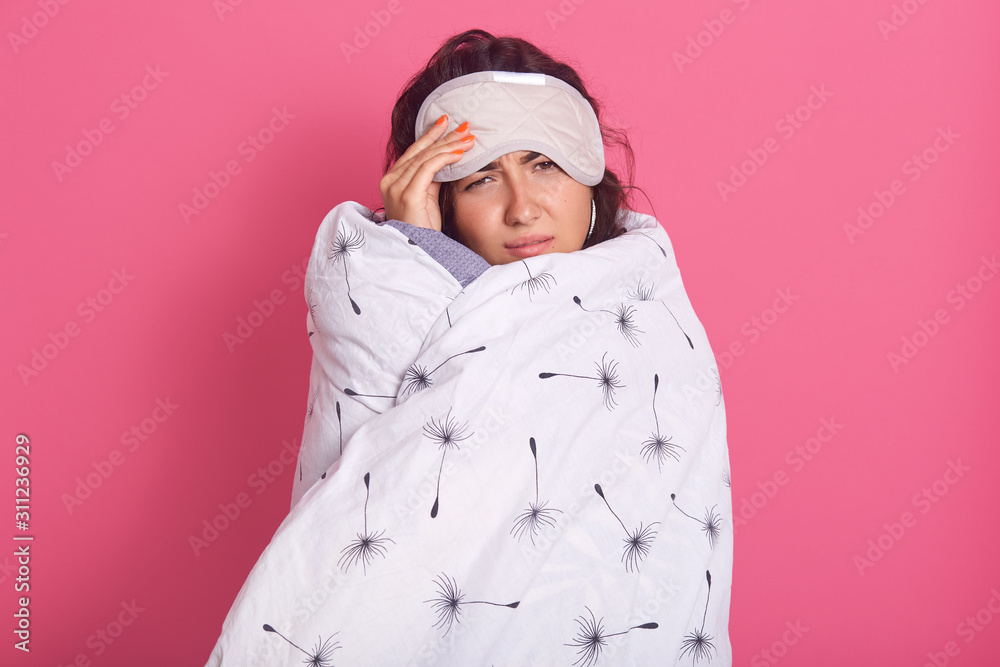 Portrait of upset woman wrapped with white blanket, female looking directly at camera, looks sad and upset, posing isolated over pink studio background, sleepy girl in morning. Portrait of upset woman wrapped with white blanket, female looking directly at camera, looks sad and upset, posing isolated over pink studio background, sleepy girl in morning.