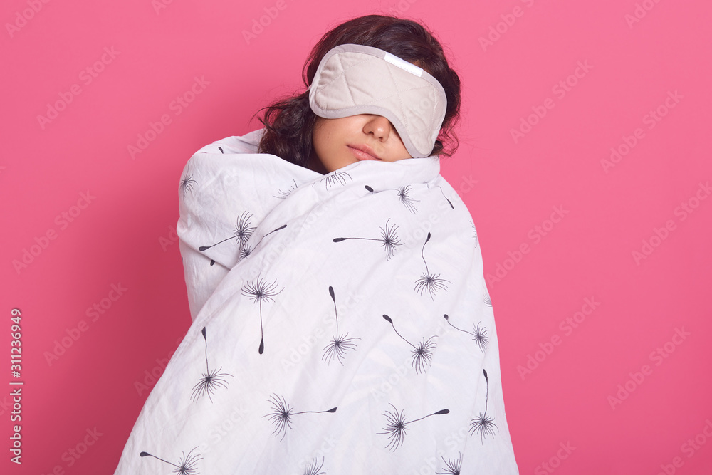 Sleep equipment concept. Portrait of brunette woman wrapped white blanket and wearing sleeping eye mask. Studio shot of young female isolated over pink background, lady being ready to fall asleep.