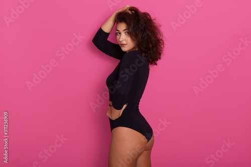 Fashion model posing isolated over pink background wearing black combi dress, standing in studio with hand one on her head and other on hip, side view of slim beautiful female with dark wavy hair.