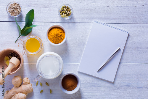 Ingredients for Golden Latte on a white wooden background. Ginger root  milk  turmeric  honey  cardamom  allspice peas and a notebook with a clean sheet.