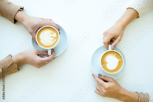 Top view of hands of two friendly females holding cups with cappuccino in cafe