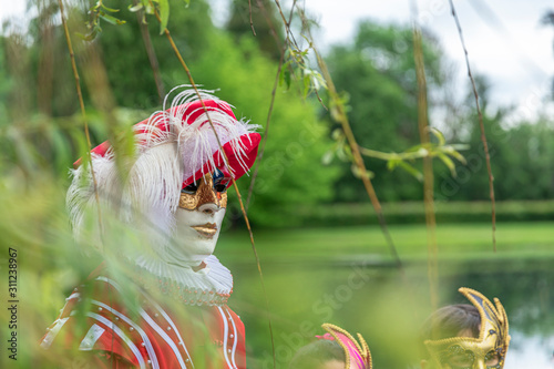 A man standing in mask and red masquerade costume agains the pond background