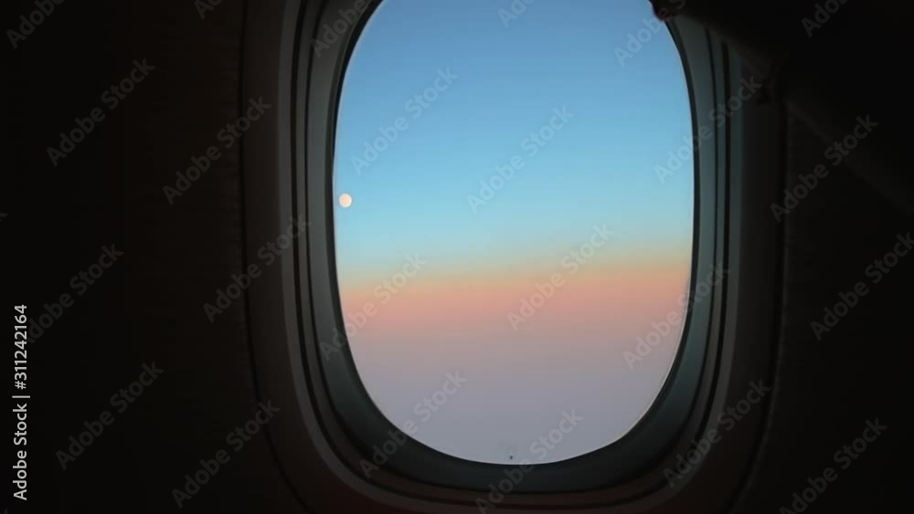 Hand closes airplane window to looks out during air travel. Passenger pov through plane window flight during sunset or sunrise. Close up of porthole of flying airplane at night. Aircraft above clouds. Hand closes airplane window to looks out during air travel. Passenger pov through plane window flight during sunset or sunrise. Close up of porthole of flying airplane at night. Aircraft above clouds.