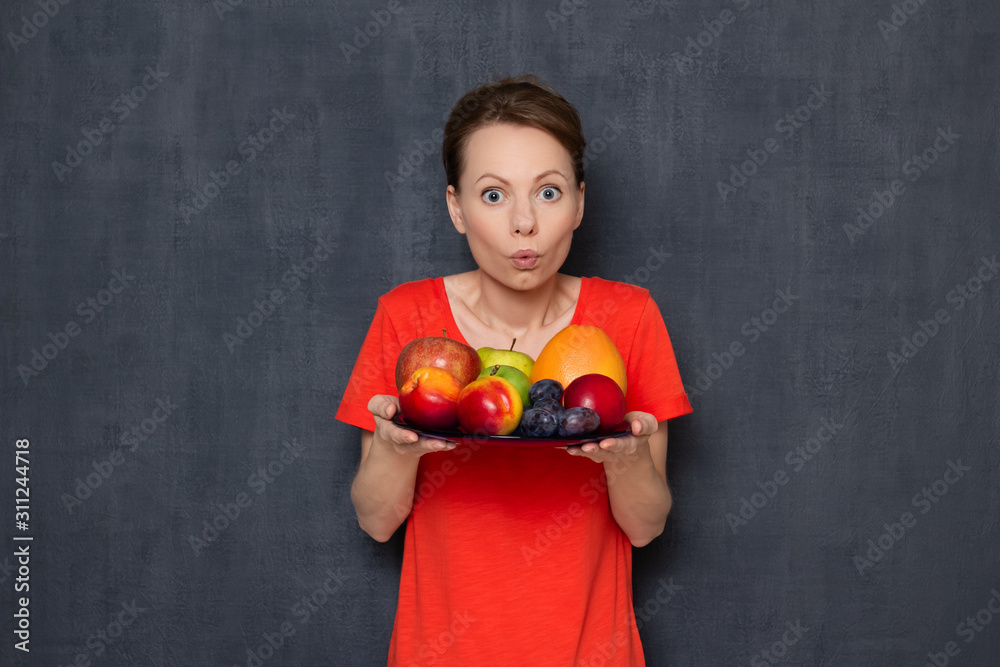 Portrait of amazed impressed young woman holding plate of ripe fruits Portrait of amazed impressed young woman holding plate of ripe fruits