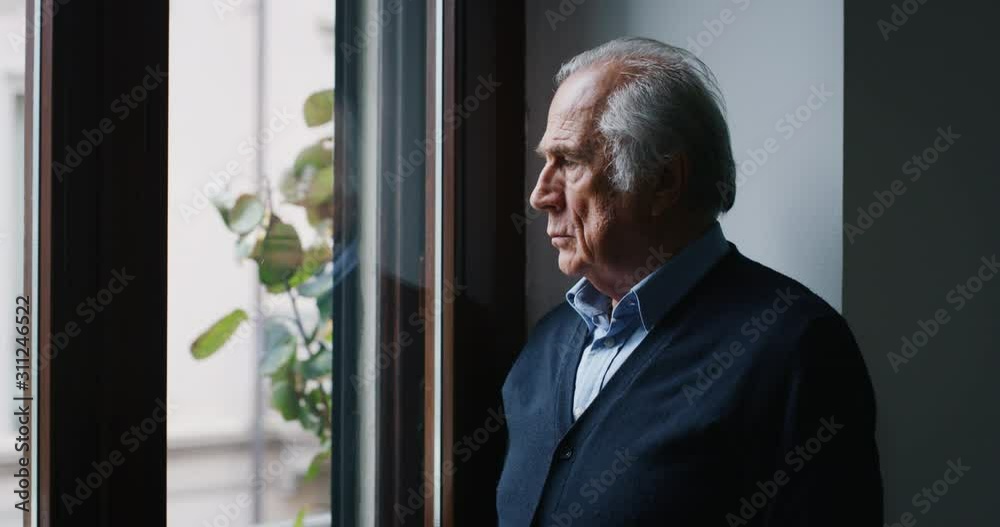 An elderly thoughtful smiling man is looking out a window of his house in the morning.