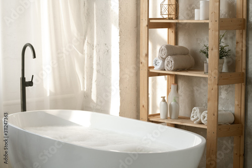 Large porcelain white bathtub filled with water and foam by wooden shelves photo