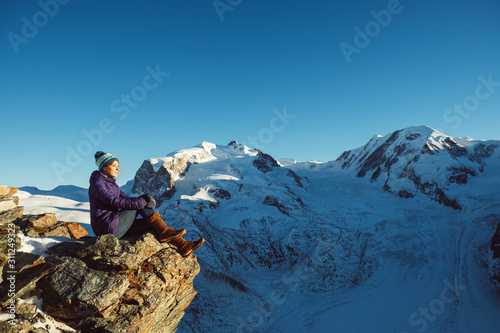 Traveler woman trekking in mountains, enjoying beautiful Swiss landscape view. Explorer girl hiking on snowy hills, travel in Alps, Switzerland. Hiker sitting on rock cliff outdoors on nature. © YOUproduction