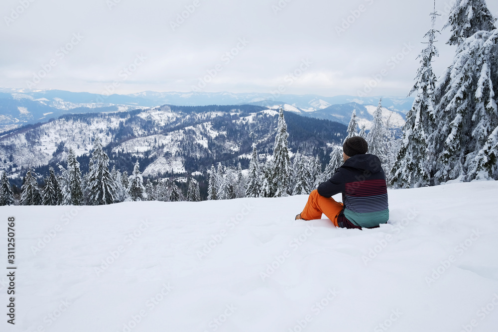 Traveler man sitting on the mountain hillside and looking on skyline view in front of beautiful pine trees in winter. Travel background with snow. Skier, explorer active holidays, vacation.