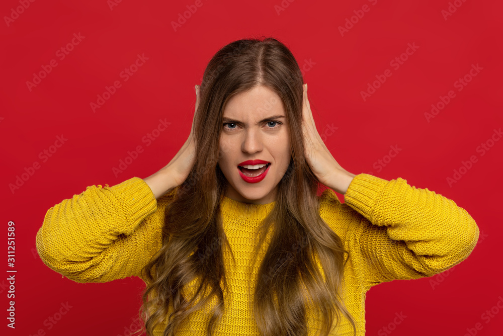 angry pretty girl in yellow sweater with red lips covering ears with hands isolated on red background. Face expressions concept