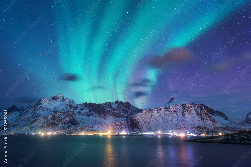 Aurora borealis over the sea, snowy mountains and city lights at night. Northern lights in Lofoten islands, Norway. Purple starry sky with polar lights. Winter landscape with aurora reflected in water