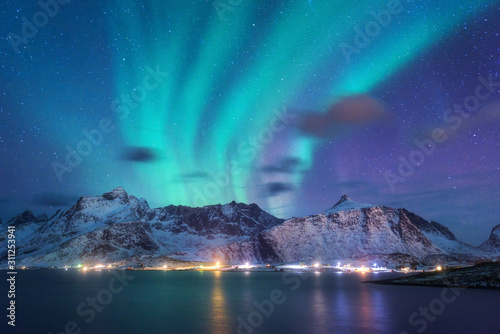 Aurora borealis over the sea, snowy mountains and city lights at night. Northern lights in Lofoten islands, Norway. Purple starry sky with polar lights. Winter landscape with aurora reflected in water © den-belitsky