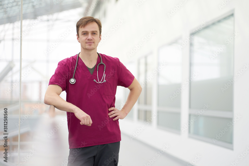 Smiling caucasian male doctor with stethoscope on neck, weared in burgundy outfit stands in white corridors of clinic
