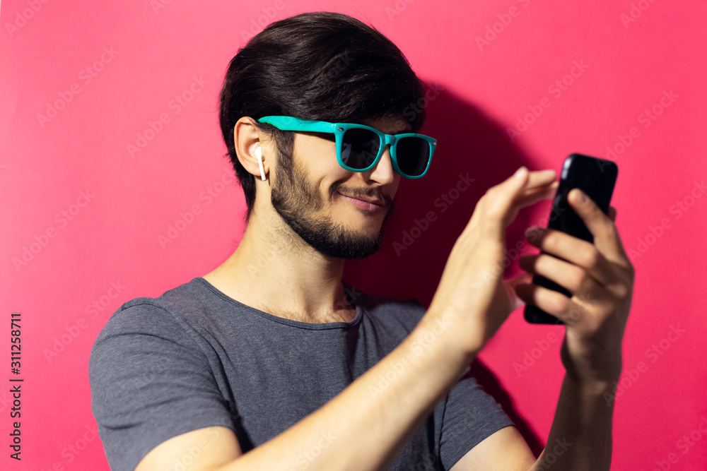 Studio portrait of young smiling man using smartphone and wireless earphones, wearing cyan sunglasses, isolated on pink coral background. Studio portrait of young smiling man using smartphone and wireless earphones, wearing cyan sunglasses, isolated on pink coral background.
