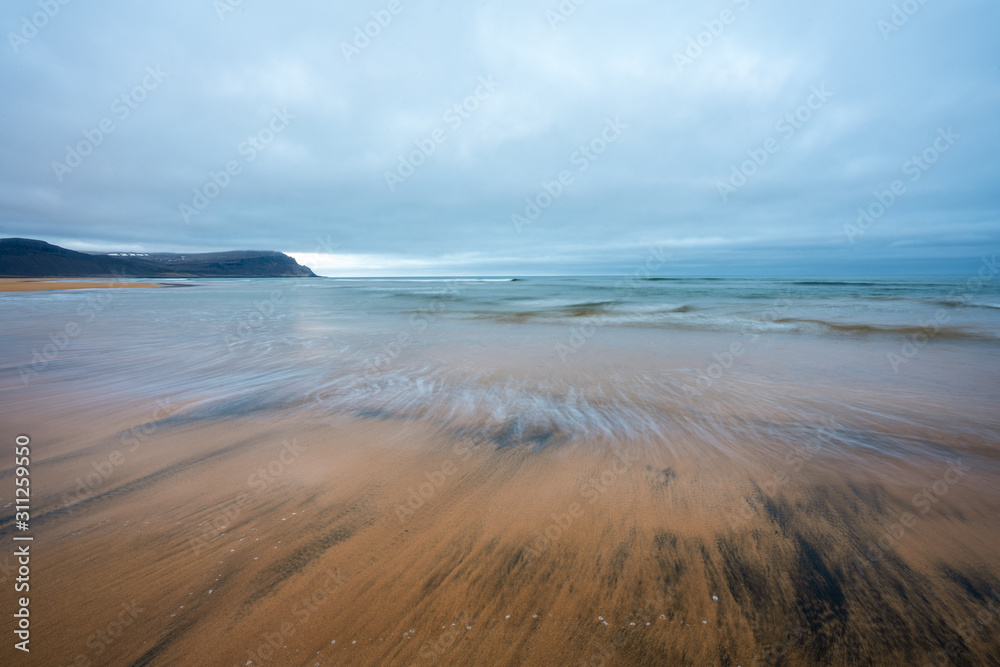 Obraz premium Raudasandur or red sandy beach in the westfjords of Iceland during blue hour. Waves crash into the red sandy dunes at low tide. Texture in the foreground and mountains in the background. 