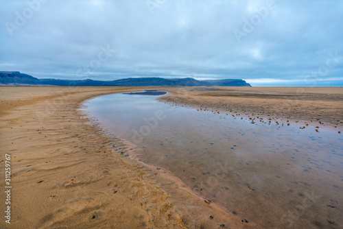 Raudasandur or red sandy beach in the westfjords of Iceland during blue hour. Waves crash into the red sandy dunes at low tide. Texture in the foreground and mountains in the background. 