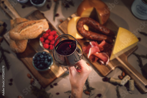 Holding a glass of red wine and in the background a table with gourmet ingredients, variety of cheeses, cold meats, grapes, raspberry, blackberry and artisanal bread on a rustic background