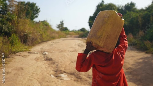 Slowmotion shot of African kid carrying water bottle on his head after fetching water from the river. Stabilize shot of walking with water bucket.