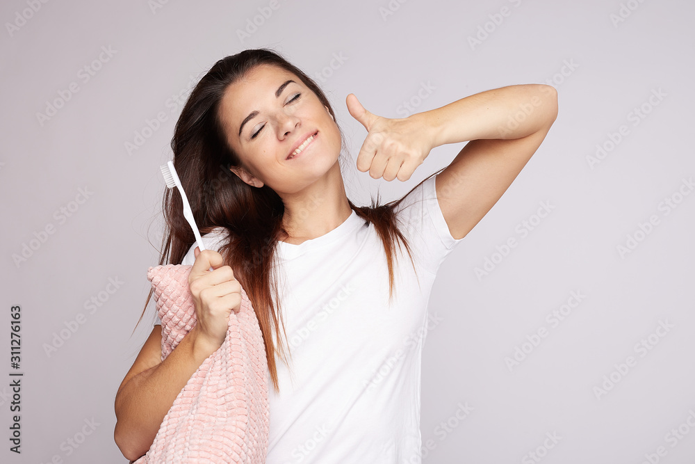 Smiling pleased young woman with closed eyes, refreshes her complexion with morning hygiene, holds brush, bites lower lip and shows white teeth, stands indoors on grey.  Body care and beauty concept.