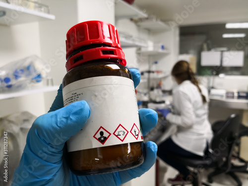 Amber bottle for protection of a hazardous chemical with GHS symbols that indicate toxicity, corrosivity and contamination. Background of a scientific laboratory with a woman doing research.