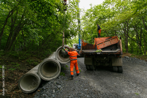 Workers in orange uniform unload drain pipes on a forest road in summer © alexhitrov