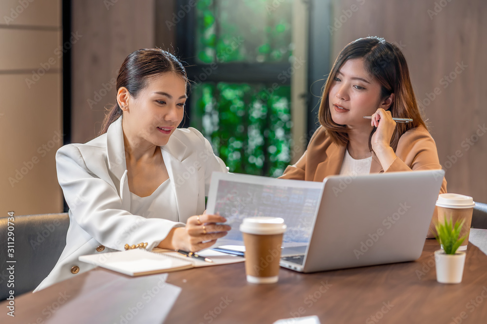 Two asian businesswomen working with the partner business with paper graph document and technology laptop in modern meeting room, office or working space, coffee break, partner and colleague concept Two asian businesswomen working with the partner business with paper graph document and technology laptop in modern meeting room, office or working space, coffee break, partner and colleague concept