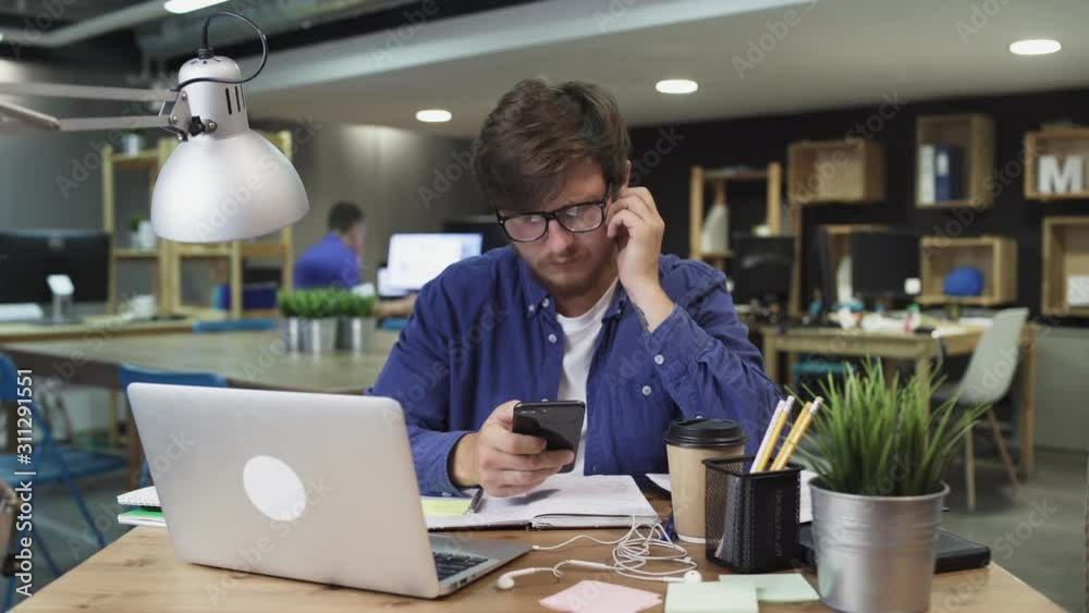 Businessman Sitting at His Desk Works on Desktop Computer in the Stylish Office, Picks up and Starts Using Smartphone, Uses Social Media App, Emailing Business Partners, Messaging. Businessman Sitting at His Desk Works on Desktop Computer in the Stylish Office, Picks up and Starts Using Smartphone, Uses Social Media App, Emailing Business Partners, Messaging.