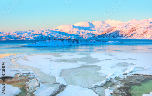 Beautiful winter lanscape with cracks on the surface of the turquoise (green) ice next to snowy mountains - Fjord and Frozen lake, Norway