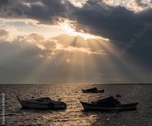 Light seeping through the clouds at sunset. Silhouettes of boats in reverse l...