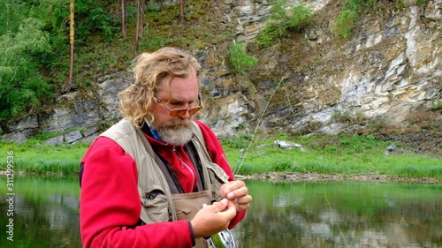 Fly fishing. A fisherman with a fishing rod ties a fly on a hook and bites the fishing line with his teeth. Hands of an elderly man close-up. Sport and hobbies.
